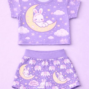 May include: A two-piece pajama set in a soft purple hue. The short-sleeved top and shorts feature a repeating pattern of a bunny on a crescent moon, stars, clouds, and elephants. The shorts have the text "Sweet Dreams".