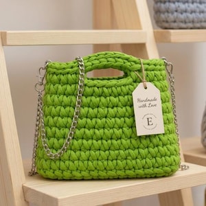 Op de afbeelding: Een felgroene, vierkante gehaakte handtas met een zilveren kettingriem en handvat. Een label met de tekst "Handmade with Love" is bevestigd. De tas wordt op een houten plank getoond.