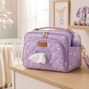 Könnte beinhalten: Eine gesteppte lavendelfarbene Wickeltasche mit Blumenmuster, ausgestattet mit einem Tragegriff, goldfarbenen Reißverschlüssen und einer Vordertasche mit Kunststoffspender für Feuchttücher. Eine Babyflasche steckt in der Seitentasche und eine passende Handschlaufe ist seitlich mit einem goldfarbenen Metallclip befestigt.