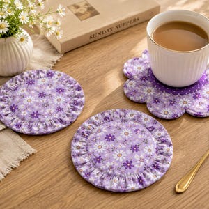 Puede incluir: Tres posavasos de tela redondos con un estampado floral morado de margaritas blancas y amarillas. Dos posavasos tienen un borde fruncido, mientras que uno tiene forma de flor. Una taza de cerámica blanca con café descansa sobre el posavasos floral.