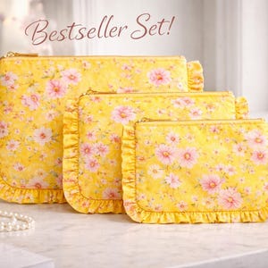 May include: A set of three yellow floral print cosmetic bags with ruffled edges. The bags feature a pink and white flower pattern and gold zippers. The largest bag has a rectangular shape, while the smallest has a wrist strap. The text "Bestseller Set!" is visible.