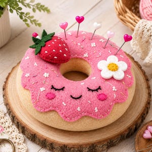 May include: A felt donut pincushion with a beige base and pink frosting, decorated with a red strawberry, a white flower, and small white embroidered details. The donut features a stitched smiling face with closed eyes and pink cheeks, and includes several heart-topped sewing pins inserted into the top.