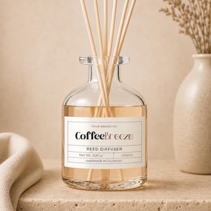 May include: A clear glass reed diffuser with wooden reeds. The label reads "CoffeeBreeze" and "REED DIFFUSER". The liquid inside is light brown. The diffuser is on a stone surface, with a vase and fabric in the background.