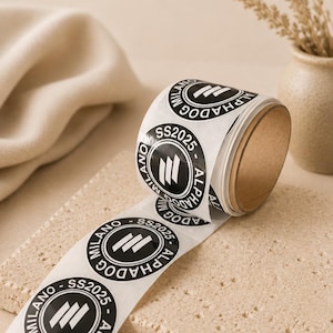 May include: A roll of white stickers with a black circular design. The design includes the text "MILANO", "ALPHADOG", and "SS2025". The stickers are on a beige surface.