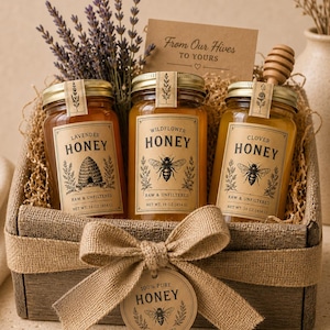 May include: A wooden gift basket filled with three glass jars of honey, labeled Lavender, Wildflower, and Clover. Each jar has a gold lid and a label with a bee illustration. A burlap bow and a sprig of lavender complete the presentation.