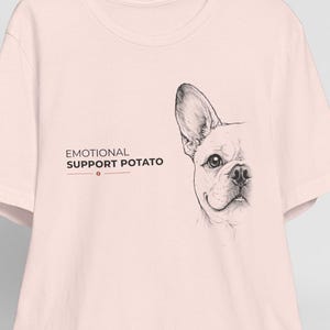 Könnte beinhalten: Ein hellrosa kurzärmeliges T-Shirt mit einer schwarzen Strichzeichnung eines Französischen Bulldoggen-Gesichts auf der rechten Seite und dem in Schwarz gedruckten Text "EMOTIONAL SUPPORT POTATO" auf der linken Seite, an einem Holzkleiderbügel.