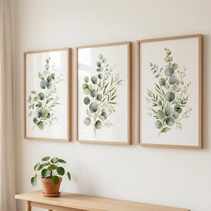 May include: Three framed botanical prints featuring watercolor eucalyptus leaves in shades of green and blue-green. The prints are in light wood frames and hang on a white wall. A small potted plant sits on a wooden bench below.