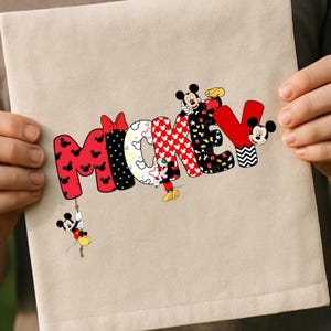May include: A beige fabric item featuring the word "MICKEY" in colorful, patterned letters. The letters are decorated with Mickey Mouse imagery, including his iconic ears and bow. A small Mickey Mouse figure hangs from the letter "M".