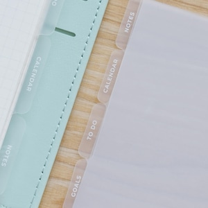 May include: A set of translucent plastic planner dividers with white text labels including Notes, Calendar, To Do, and Goals. These dividers are positioned alongside a light mint green planner cover featuring stitched edges and grid paper.