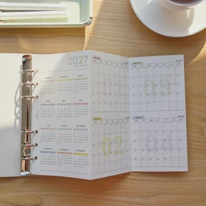 Peut inclure: Un agenda annuel 2027 avec un mécanisme de reliure à six anneaux en métal argenté. Les pages ouvertes présentent une grille de calendrier annuel avec des sections mensuelles, des en-têtes colorés et de grands chiffres translucides pour les mois.