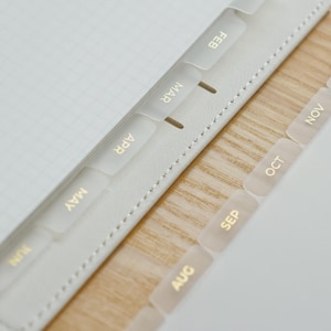 May include: A white planner with grid paper pages and translucent dividers featuring gold foil text for the months of February, March, April, May, June, August, September, October, and November, with visible white stitching along the edge.