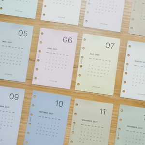 May include: A collection of pastel-coloured monthly calendar pages for the year 2027, featuring months May through December. Each rectangular paper sheet includes a large month number, the month name, a grid of dates, and the text LET'S PLAN.