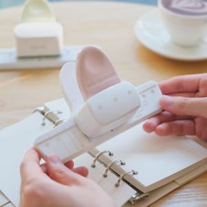May include: A white and light pink paper punch tool with the text 4MM ORGANIZER printed on the top. The device features a horizontal guide bar with measurement markings and is positioned over a grid-lined paper insert for a ring-bound planner.