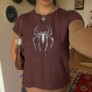 May include: A maroon short-sleeved t-shirt with a white spider graphic. The spider design is centered on the front. The shirt is made of a soft material.