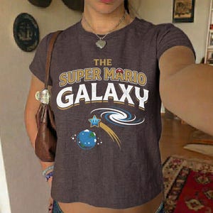 May include: A brown t-shirt featuring the text "THE SUPER MARIO GALAXY" in gold and white. The shirt has a graphic of a planet, a star, and a galaxy swirl. The shirt is short-sleeved.