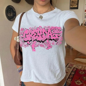 May include: White t-shirt with a pink graffiti-style design that reads "GREYDAY 2026". The design features stylized lettering and illustrations of figures. The shirt is short-sleeved and appears to be made of cotton.