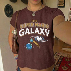 May include: A maroon t-shirt featuring the text "THE SUPER MARIO GALAXY" in white and gold lettering. Below the text is a graphic of a blue planet, a shooting star, and a spiral galaxy design.