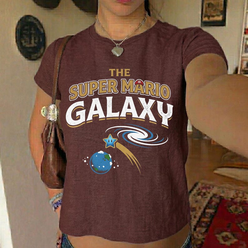 May include: A maroon t-shirt featuring the text "THE SUPER MARIO GALAXY" in white and gold lettering. Below the text is a graphic of a blue planet, a shooting star, and a spiral galaxy design.
