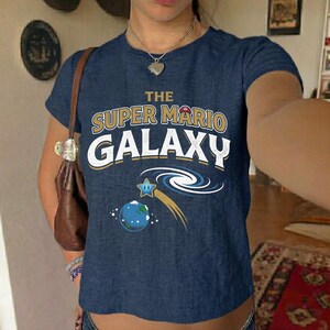 May include: A navy blue t-shirt featuring the text "THE SUPER MARIO GALAXY" in gold and white. Below the text is a graphic of a blue planet, a shooting star, and a swirl design. The shirt has short sleeves.