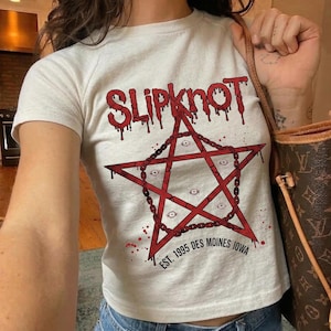 May include: A white t-shirt featuring a red pentagram design with a chain border and multiple eye graphics. The word "Slipknot" is printed above the pentagram in a dripping font. The text "EST. 1995 DES MOINES IOWA" is below the pentagram.