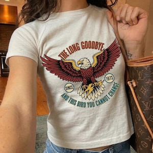 May include: Cream-colored t-shirt featuring a graphic of a bald eagle with outstretched wings. The eagle is in shades of red, yellow, and white, with a lightning bolt in its talons. Text includes "THE LONG GOODBYE" and "AND THIS BIRD YOU CANNOT CHANGE."