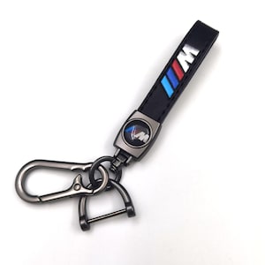 May include: Black leather keychain with a metal clasp and a silver-tone metal accent. The keychain features the BMW M logo in blue, red, and white stripes. The keychain is designed for car keys.
