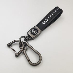 May include: Black leather keychain with the word "INFINITI" in white lettering. The keychain features a silver-colored metal clasp and a metal emblem with the Infiniti logo. The keychain is designed for car keys.