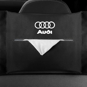 May include: Black car tissue holder featuring the Audi logo in white. The rectangular holder has a dispensing slot and is attached to a car seat headrest.