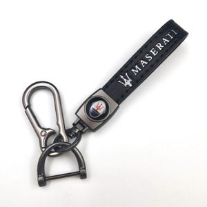 May include: Black leather keychain with the word "MASERATI" in white lettering. The keychain features a metal clasp and a circular emblem with the Maserati logo in the center.