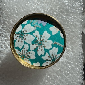 Może przedstawiać: Okrągły pojemnik ze złotą obwódką i turkusowym wzorem kwiatowym. Wzór przedstawia białe kwiaty ze złotymi akcentami. Pojemnik leży na folii bąbelkowej.