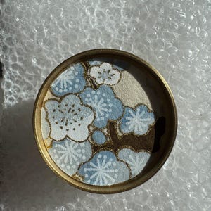 May include: A round, decorative coaster with a gold rim. The coaster features a floral design in shades of blue, white, and gold. The flowers are stylized and arranged in a repeating pattern. The coaster is likely made of a durable material.