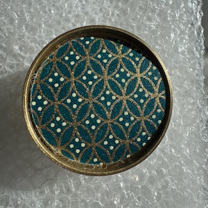 Può includere: Contenitore rotondo con un motivo geometrico verde acqua e oro. Il contenitore ha un bordo dorato e un interno verde acqua con un motivo ripetuto oro e bianco. È appoggiato su pluriball.