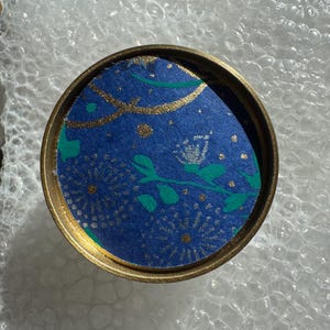 Può includere: Un contenitore rotondo con bordo dorato e interno blu. L'interno presenta un design con dettagli dorati e argentati, insieme a forme di foglie verdi. Il contenitore è appoggiato su pluriball.