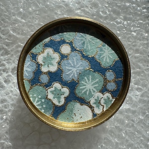 May include: A round, gold-colored container with a floral design. The interior features a blue background with light blue, white, and green flower patterns. Gold accents are visible throughout the design. The container is likely a decorative item or a small storage box.