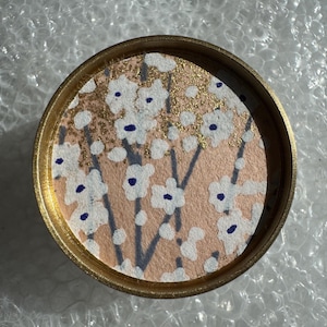 以下が含まれることがあります： 金色の縁取りと花柄が特徴の丸いコースター。 桃色の背景に白い花、ネイビーブルーの中心、金色のアクセントが施されています。 花は濃い灰色の茎に配置されています。