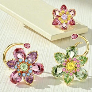 May include: Jewelry set featuring flower-shaped rings and earrings. The rings and earrings have a gold-tone metal base and are adorned with pink, green, yellow, and blue crystals. The earrings have a curved design.