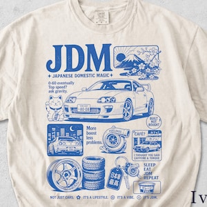 Könnte beinhalten: Elfenbeinfarbenes T-Shirt mit blauem Grafikdesign, das ein Auto, japanischen Text und die Buchstaben "JDM" zeigt. Das Design umfasst eine Glückskatze, eine Berglandschaft und Autoteile. Das Shirt hat einen Rundhalsausschnitt und kurze Ärmel.