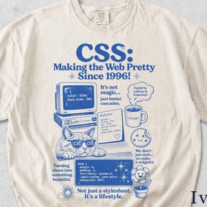 Op de afbeelding: Ivoor kleurig t-shirt met een blauw grafisch ontwerp. Het ontwerp bevat de tekst "CSS: Making the Web Pretty Since 1996!" samen met illustraties van een computer, een kat met zonnebril, een koffiekopje en een koekje.
