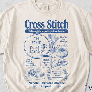 May include: Ivory t-shirt featuring a blue cross-stitch design. The design includes the words "Cross Stitch" and "Stabbing fabric politely since forever." Other elements include a cat in a hoop, scissors, and a coffee mug with text.