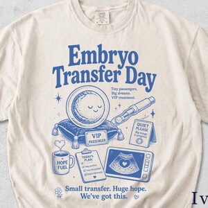 Könnte beinhalten: Ein elfenbeinfarbenes Kurzarm-T-Shirt mit blauen grafischen Illustrationen und Text. Das Design zeigt die Worte Embryo Transfer Day, eine lächelnde kreisförmige Figur auf einem Kissen, ein medizinisches Instrument, ein Schild mit der Aufschrift Quiet Please, eine Kaffeetasse, eine Checkliste und ein Ultraschallbild.