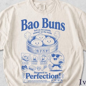 May include: Ivory t-shirt featuring a blue graphic of bao buns in a steamer, with the text "Bao Buns" and illustrations. The design includes the text "Soft on the outside, steamin' on the inside" and "Steamed to Perfection!"
