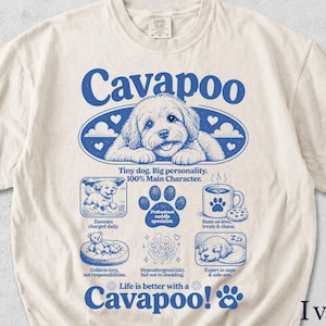 Könnte beinhalten: Elfenbeinfarbenes T-Shirt mit blauem Cavapoo-Hunde-Motiv. Das Design enthält das Wort "Cavapoo" sowie weitere Texte und Icons zum Thema Hund. Das T-Shirt ist aus weichem Material.