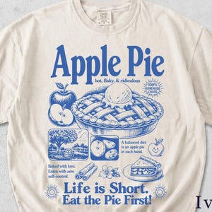 Könnte beinhalten: Cremefarbenes T-Shirt mit blauem Grafikdesign. Das Design enthält die Worte "Apple Pie" und Illustrationen von Apfelkuchen, Äpfeln und anderen verwandten Bildern. Der Text "Life is Short. Eat the Pie First!" ist ebenfalls enthalten.