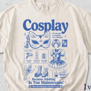 Könnte beinhalten: Elfenbeinfarbenes T-Shirt mit blauem Grafikdesign. Das Design zeigt das Wort "Cosplay", Illustrationen von Cosplay-Artikeln und den Text "Because Adulting Is Too Mainstream!" in Blau.