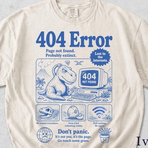 May include: Ivory-colored t-shirt with a blue graphic design. The design features a dinosaur, a computer screen displaying "404 NOT FOUND," and text that reads "404 Error" and "Don't panic." The shirt has a crew neck and short sleeves.