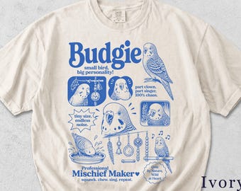 Può includere: T-shirt color avorio con un design grafico blu con la parola "Budgie" e illustrazioni di cocorite. Il design include il testo "small bird, big personality!" e "Professional Mischief Maker."