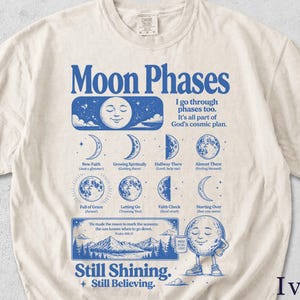 May include: An ivory short-sleeve t-shirt featuring a blue graphic print of moon phases, celestial illustrations, and text including "Moon Phases," "I go through phases too," and "Still Shining. Still Believing." with mountain landscape imagery.