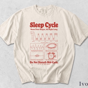 May include: An ivory short-sleeve t-shirt featuring red graphic text and illustrations detailing the sleep cycle. The design includes a sleep stage graph, an eye, a brainwave pattern, and an alarm clock with the text Sleep Cycle, Same Four Stages, All Night Long, and Do Not Disturb Mid-Cycle.