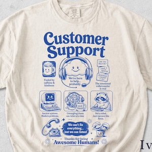 Könnte beinhalten: Elfenbeinfarbenes T-Shirt mit blauem Grafikdesign. Das Design enthält die Worte "Customer Support" und Illustrationen wie eine Kaffeetasse, eine Wolke mit Kopfhörern, einen Computer, einen Waschbären und ein Telefon. Text enthält "Fueled by caffeine & kindness."