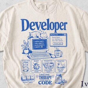 Pode incluir: Camiseta cor marfim com um design gráfico azul. O design inclui a palavra "Developer", um gato dormindo, um computador e vários ícones relacionados à codificação e ao desenvolvimento de software. O texto "I don't need therapy, I just need to code" também está incluído.